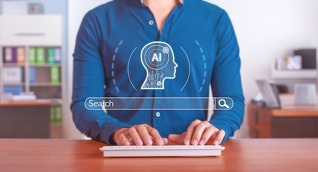 A person in a long-sleeved blue collared shirt sits at a wooden desk, typing on a white keyboard in this medium shot. In front of the person is a graphic of a search bar with the word "search" in it. Above that is a line drawing of a head with AI inside of it.