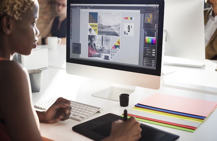 A woman with short, light-colored hair sits at a white desk in a brightly lit urban workplace, focused intently on design work. A large computer monitor displays a design in progress.