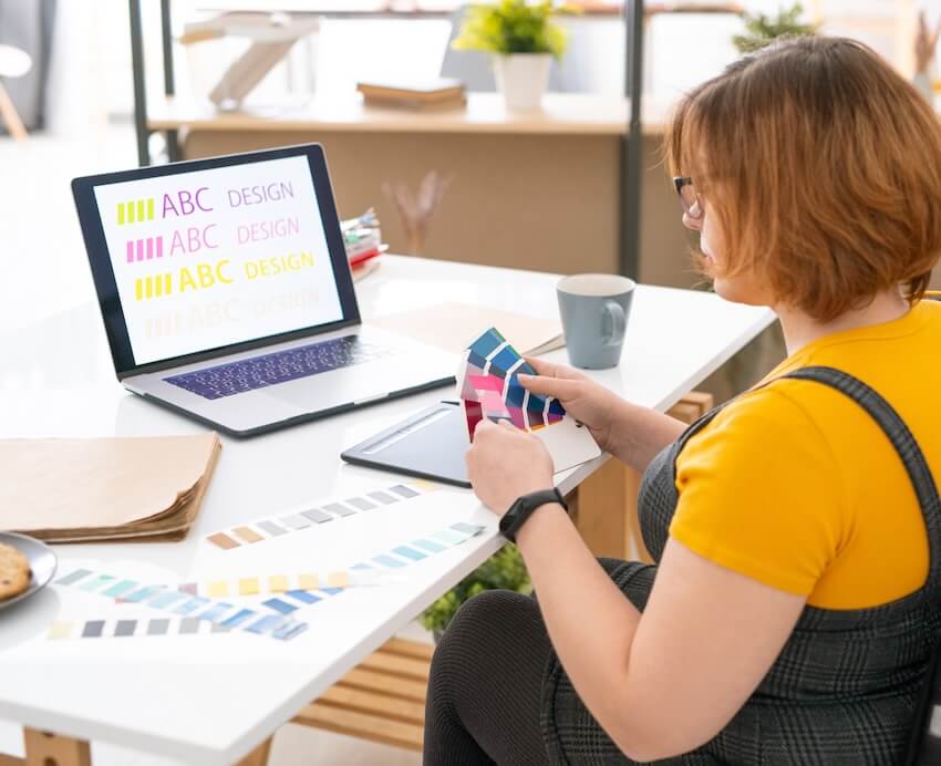 A woman with short auburn hair wearing glasses and a yellow t-shirt with a grey dress on top is seated in a wheelchair at a white desk. She is looking at a multicolored palette in her hands. On the white desk is a laptop, tablet and other design materials.