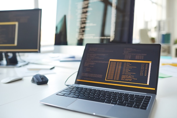 A sleek grey laptop sits open on a white desk, displaying lines of yellow code on its screen. Behind the laptop, a monitor and other office equipment are visible in the background.