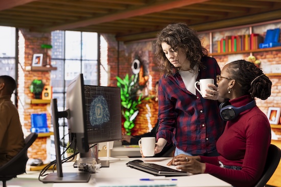 Two women in business attire are collaborating in a vibrant, contemporary office space. One woman with curly hair is standing, holding a mug and looking at a computer screen, while a second woman is sitting at a desk and holding a mug.