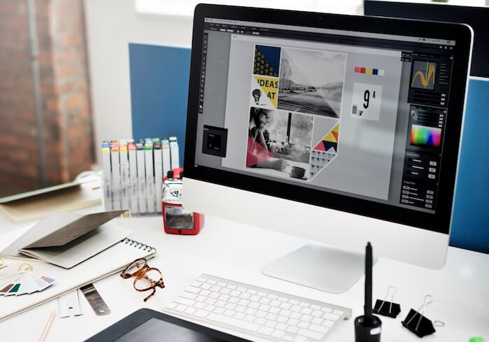 A modern computer monitor sits on a bright white desk, displaying a graphic design project with images, color palettes, and typography. The workstation includes design elements such as sketchbooks, color swatches, markers, and drawing tools.