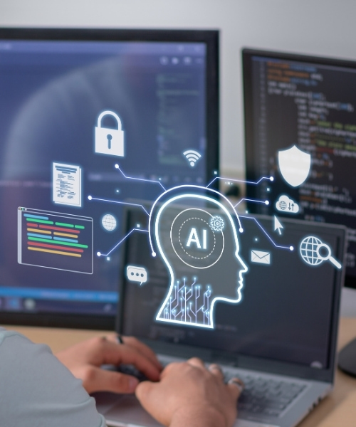 A person types at a laptop in a modern office setting. The laptop screen displays a stylized head outline with 'AI' inside, connected to icons representing technology concepts, including security and connectivity.