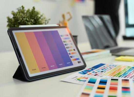 This photo captures a variety of color palettes displayed on a digital tablet, alongside physical color charts and office items on a clean white desk.