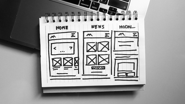 Black and white photo features a notepad containing website wireframes next to a laptop. The notepad displays hand-drawn sketches of website layouts with titles 'Home,' 'News,' and 'Modal.'