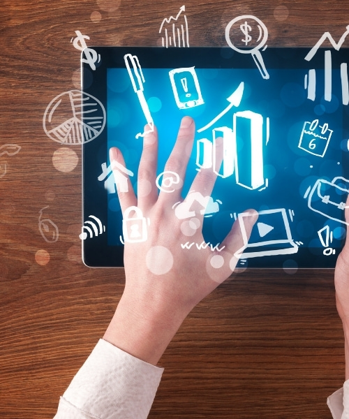 An eye-level shot shows a person's hands holding a tablet with a blue screen displaying business icons, set against a wooden background. The hand is placed on the screen to control the tablet and review the icons. The scene suggests modern business practices and technological solutions for planning and strategy.
