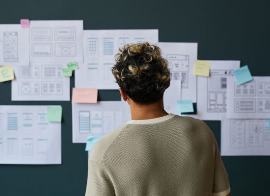 Young man with curly hair wearing a beige sweater looking at a wall with UX, website, and design elements on it.