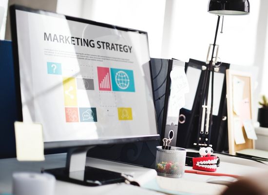 A desktop computer in an office with bright lighting. On the desktop computer screen it has charts and elements under a heading "Marketing Strategy".