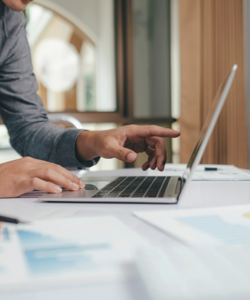 Person pointing to a laptop while working on a desk scattered with paper.