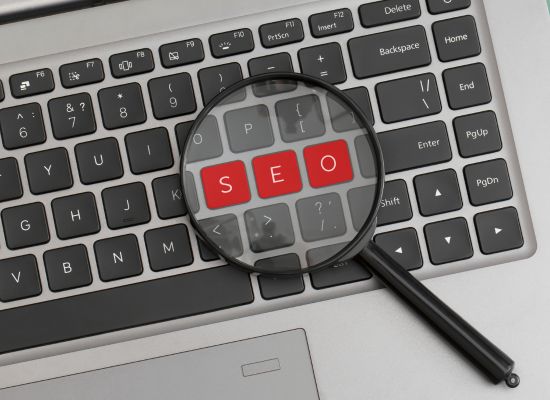 A computer keyboard fills the shot, and resting on top is a magnifying glass. The magnifying glass is positioned over keys that spell out "SEO" in red.