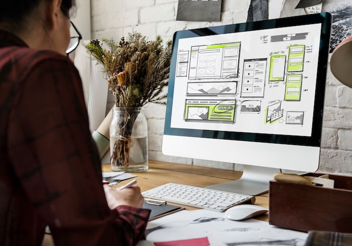An adult with dark hair wearing glasses sits at a desk with a computer. The computer displays a wireframe mockup with sketches, color blocks, and notes.