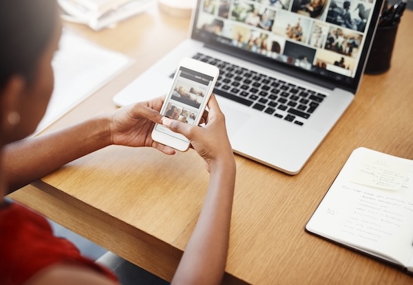 An adult sits at a wood desk using a smartphone. The desk also has an open laptop and a notebook. The laptop appears to display a collection of stock photos.