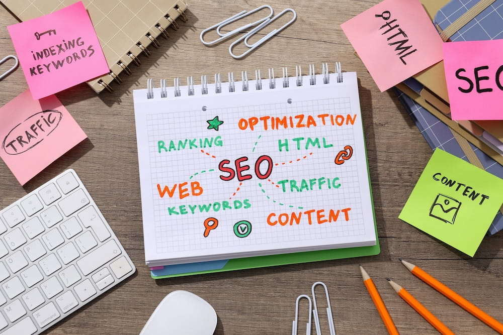This photo displays a notebook with an SEO concept drawing on a woodgrain table. Colourful sticky notes with marketing concepts like traffic and content surround the notebook, alongside a keyboard and pencils.