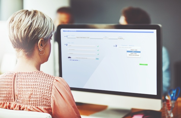 From behind, an adult works at a computer with an online form displayed. The woman has short hair and wears a patterned top. She is sitting in front of a modern desktop computer, showing an order form complete with credit card payment options.
