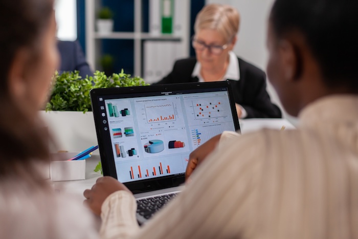 Three individuals are working in an office setting. The primary focus is on a laptop displaying business-related charts and data.