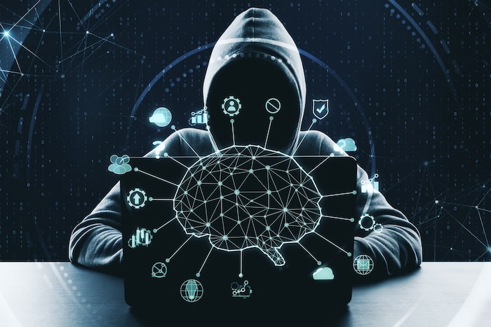 A figure wearing a hoodie sits at a table behind a laptop. The laptop screen shows an image of a brain-like neural network. Connected to the neural network are floating symbols of digital devices, like the cloud or a computer screen — signifying cybersecurity or AI and online threats.