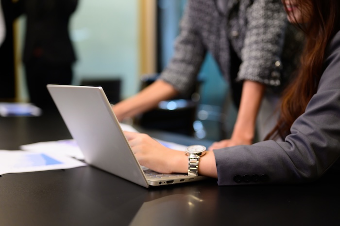 Two figures engage with a silver laptop on a black table, amidst scattered papers. The blurred figure in the background, clad in a patterned gray top, leans in, contributing to the collaborative atmosphere.