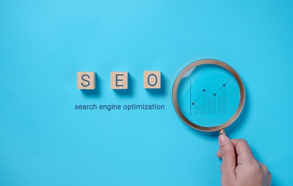 A hand is shown holding a magnifying glass over the words SEO, spelled out in wooden blocks, with the phrase "search engine optimization" written beneath.